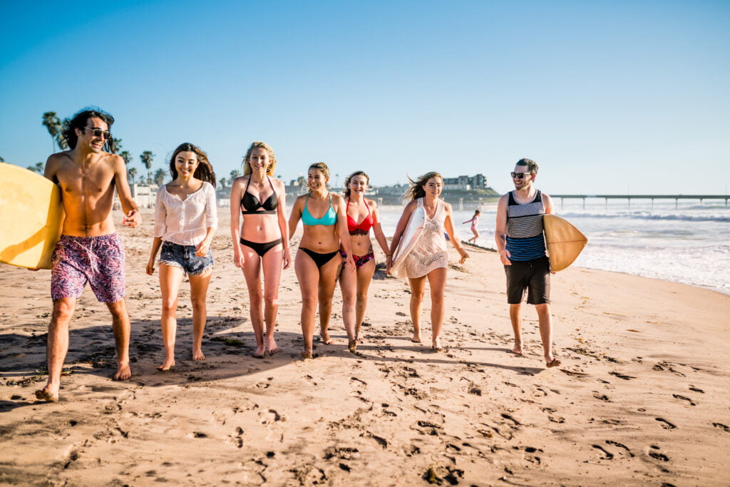 group of joyful young people dressed in swimwear, holding their surfboards, marching along the beach, laughing to each other